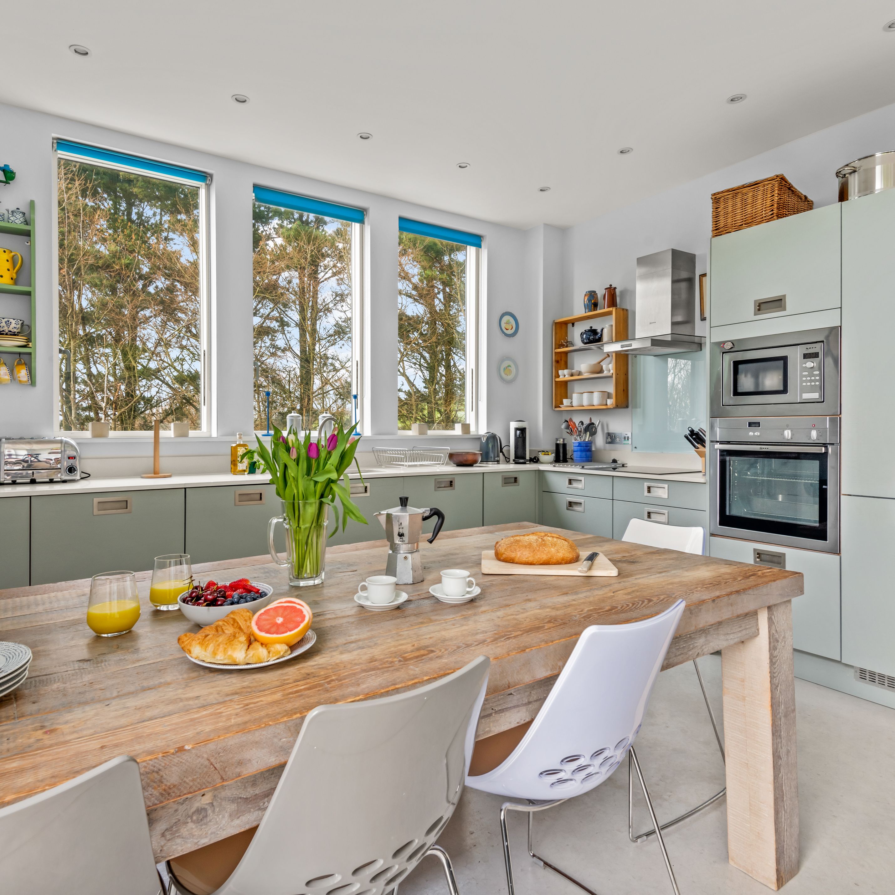 Bright modern kitchen with a rustic wooden table set for breakfast, featuring large windows, light green cabinets, and built-in appliances.