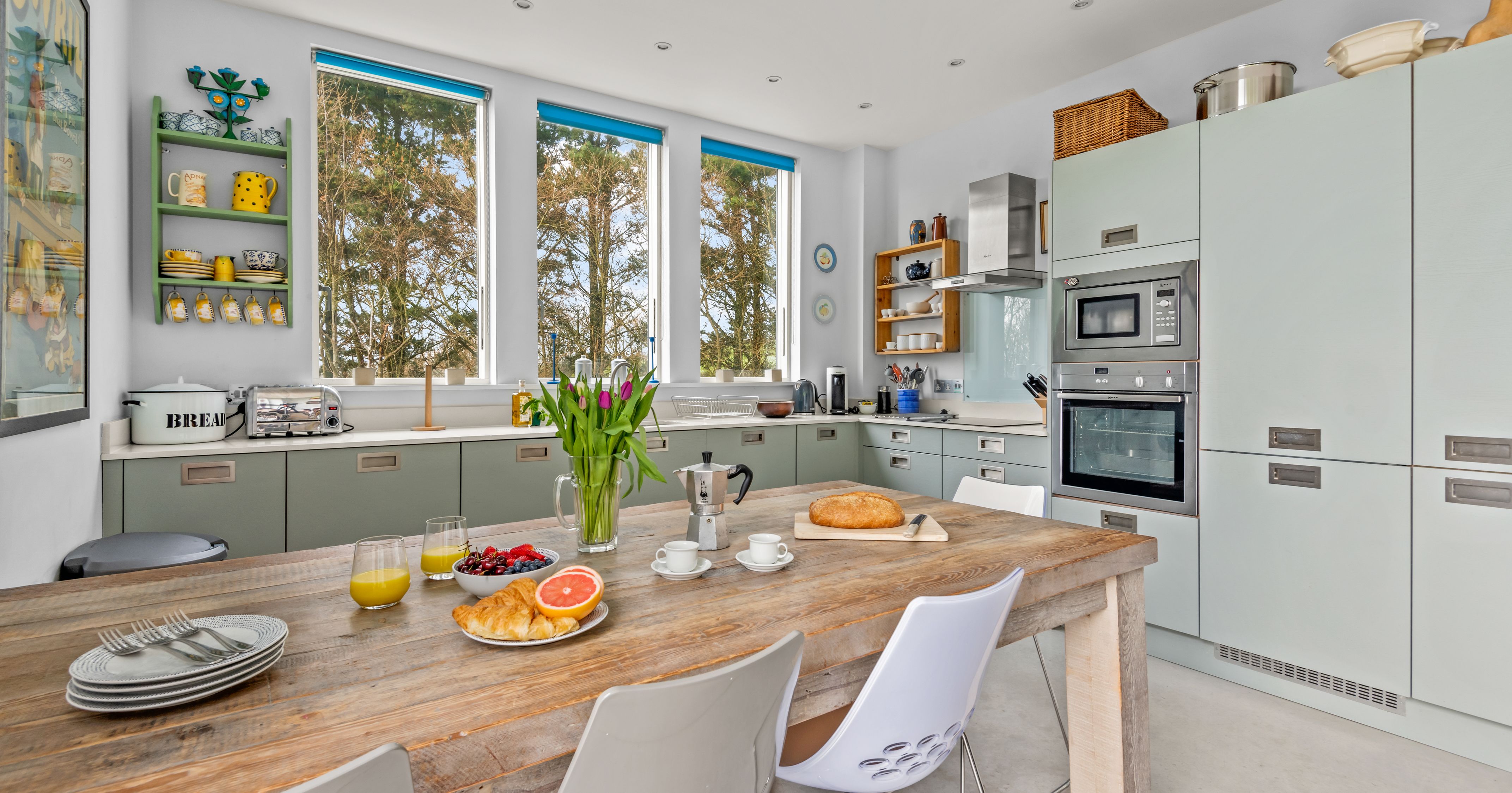 Bright modern kitchen with a rustic wooden table set for breakfast, featuring large windows, light green cabinets, and built-in appliances.