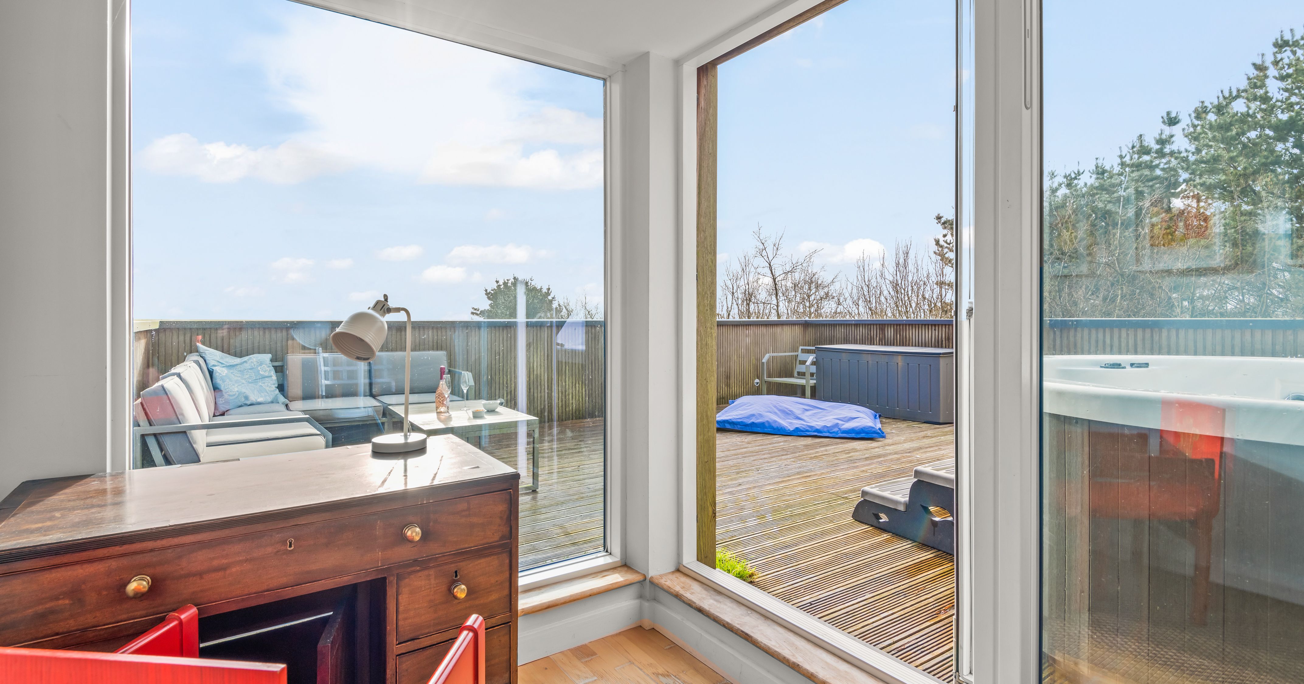 Modern home office with wooden desk and patio door leading to a spacious outdoor deck with seating, hot tub, and bean bag.