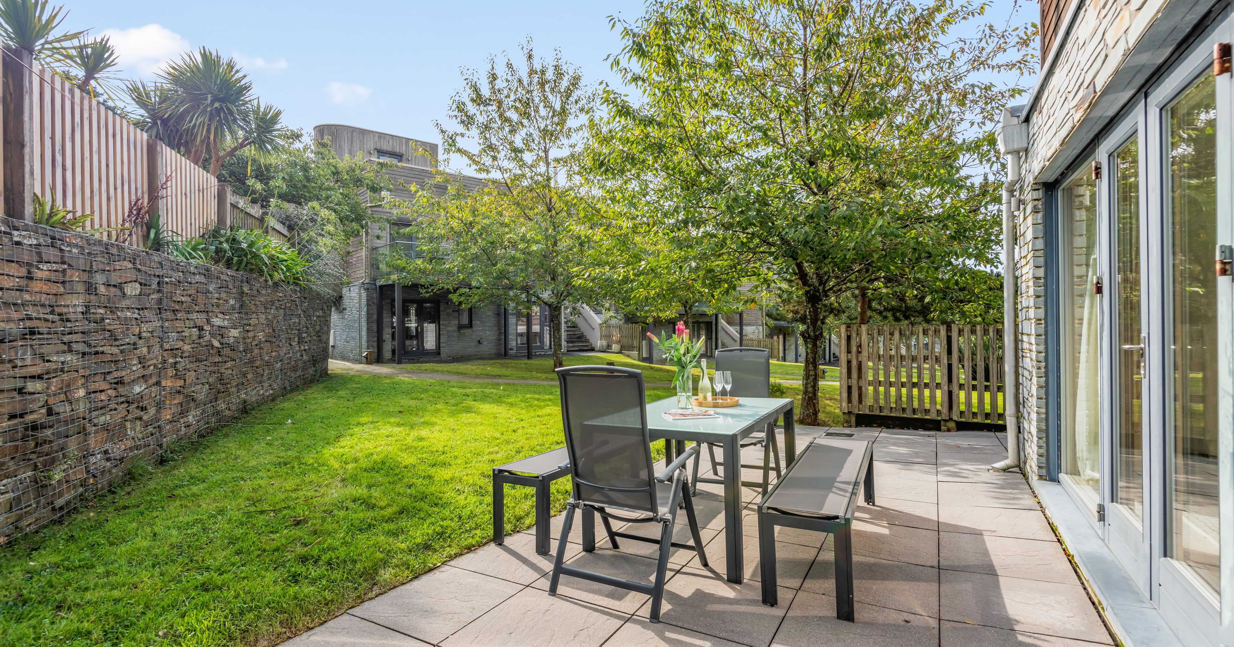 Modern outdoor patio area with a glass dining table, benches, and chairs set on a paved area next to a grassy lawn and trees, adjacent to a stone building with large windows.