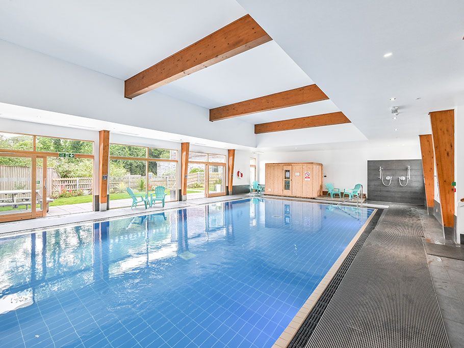 Indoor swimming pool with blue tiles, wooden beams, and large windows overlooking a garden area.