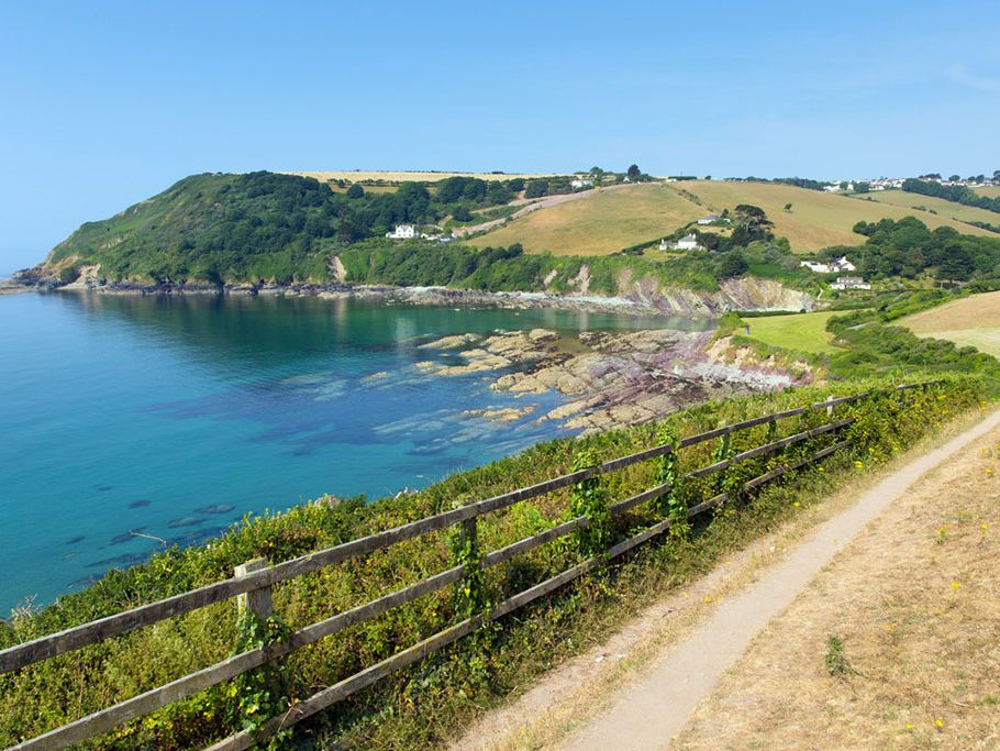 Coastal path with wooden fence overlooking a rocky bay and rolling hills with scattered houses