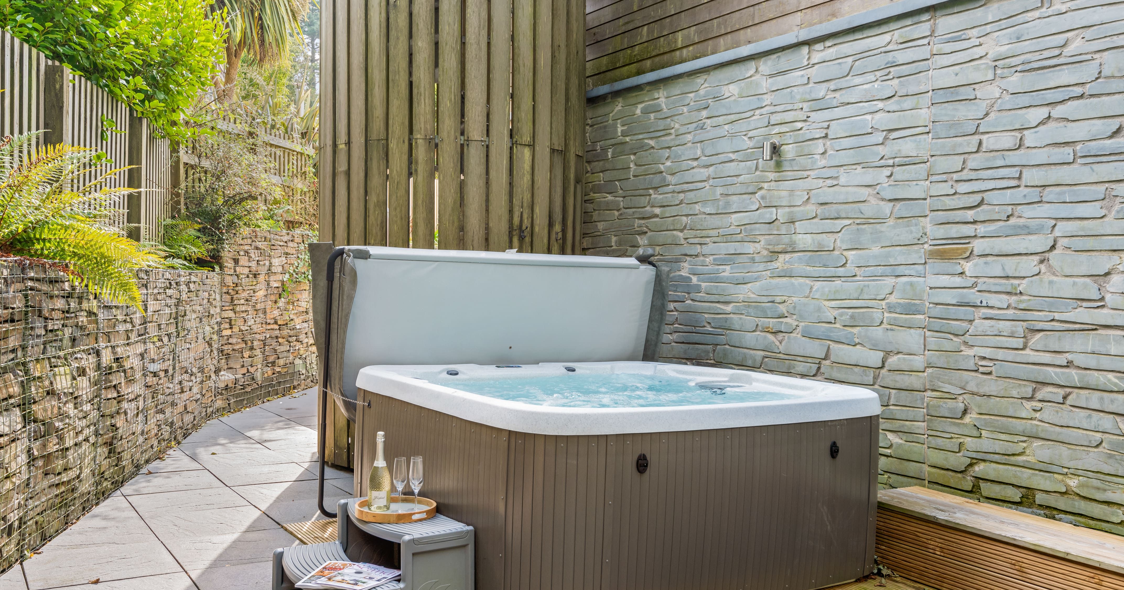 Outdoor hot tub with bubbling water, surrounded by stone and wooden walls, greenery, and a small step with a tray holding champagne and glasses.