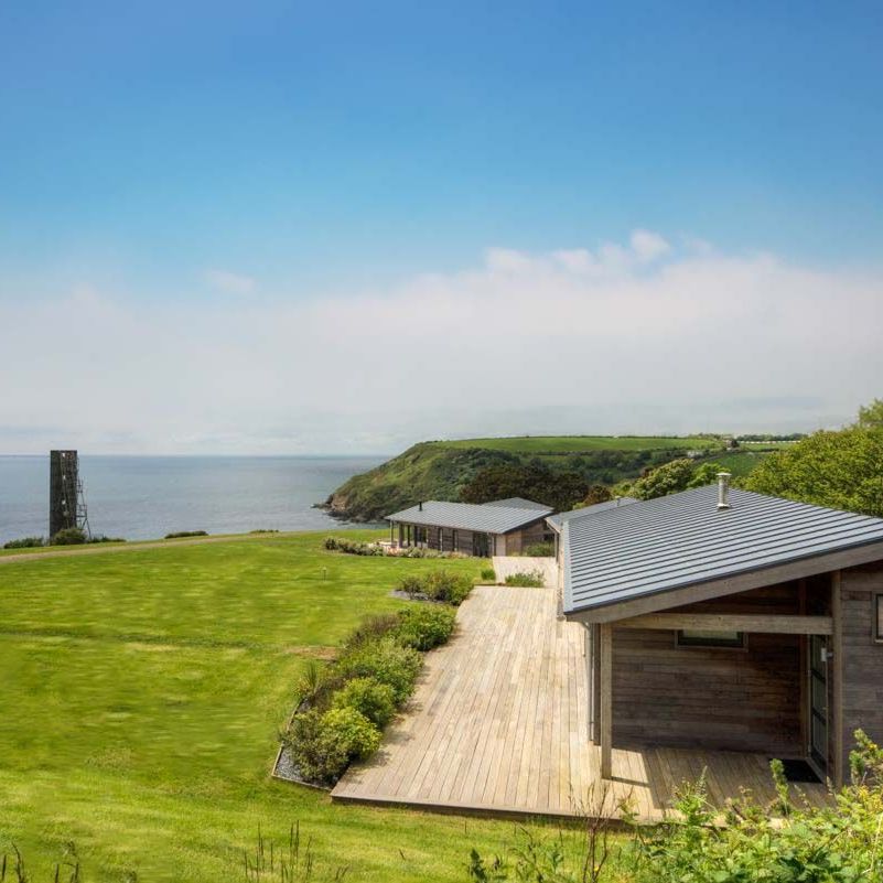 Modern cabin with a wooden deck overlooking a grassy lawn and coastline with a tower by the sea