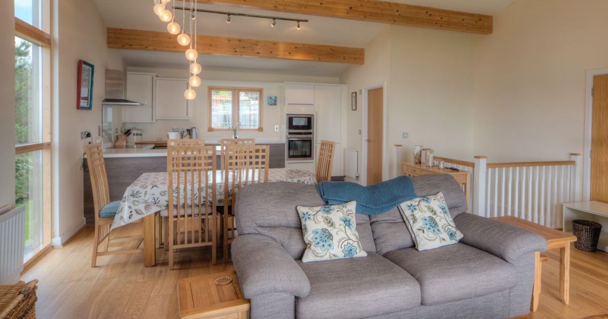 Modern open-plan living and dining area with wooden beams, grey sofa, and wooden furniture.