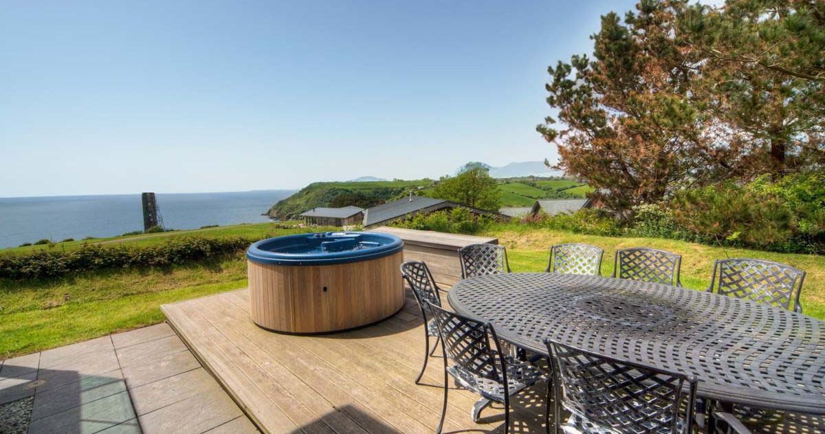 Outdoor patio with hot tub, table and chairs overlooking the sea and green hills.