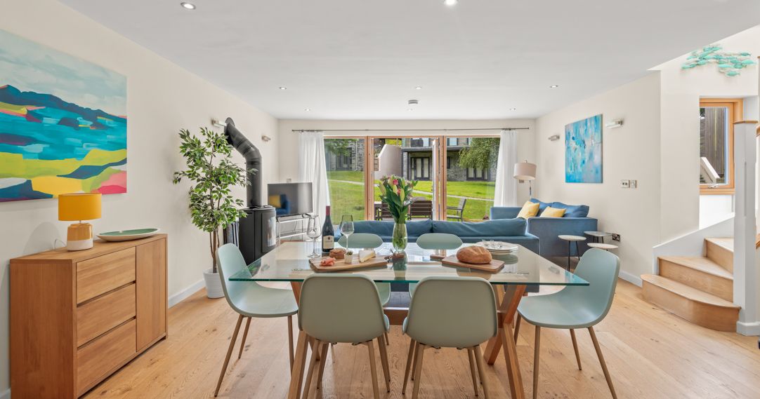 Modern open-plan living and dining area with glass dining table, light blue chairs, wooden floor, blue sofa, colorful abstract paintings, and large windows overlooking a green lawn.