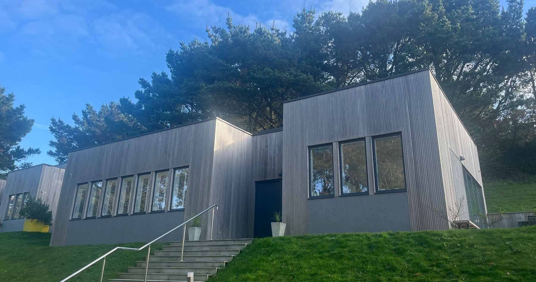 Modern rectangular building with large windows and wooden exterior, surrounded by grass and trees.