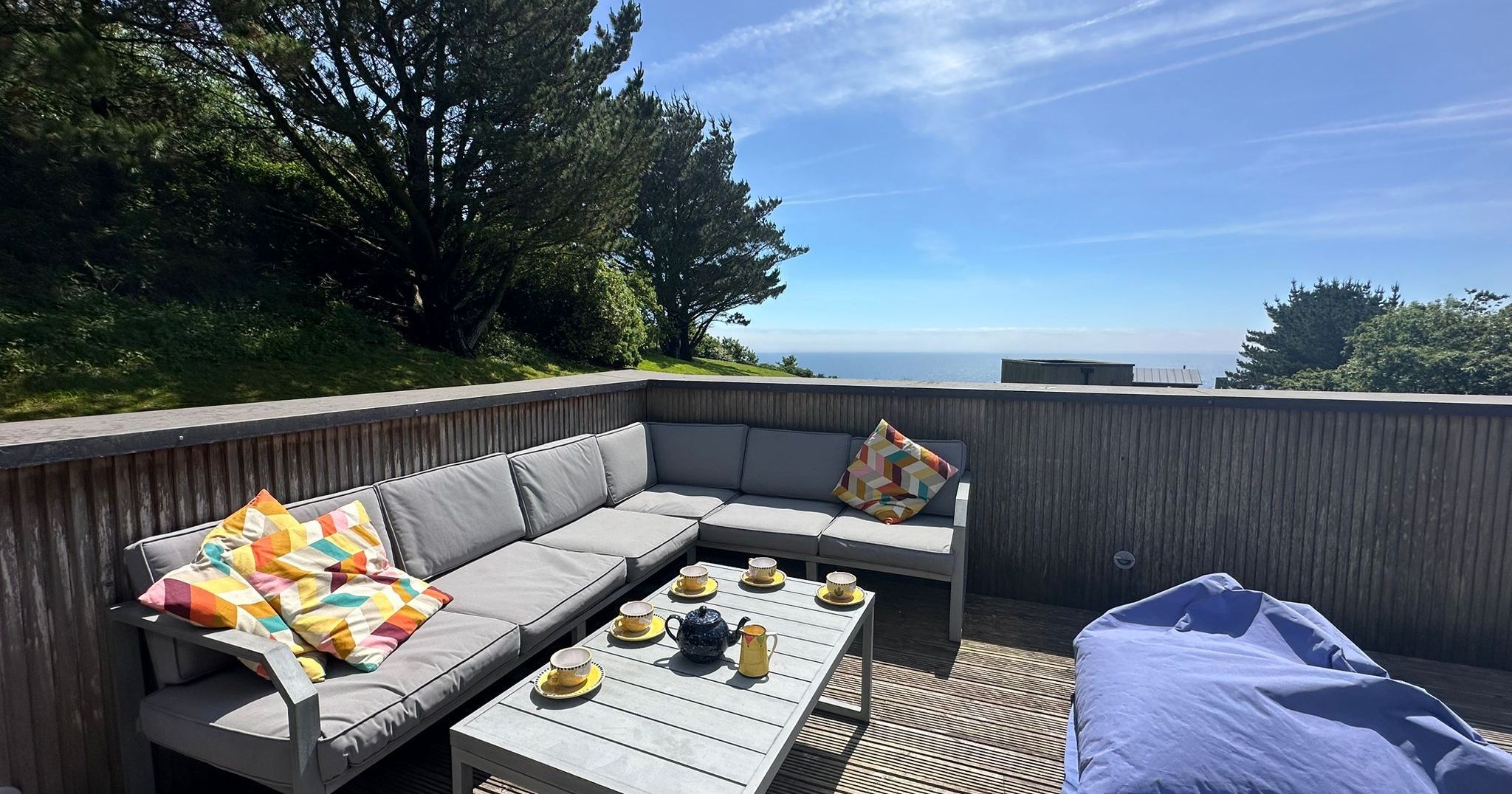 Outdoor patio with sectional sofa, beanbag, coffee table with tea set, bright cushions, and a view of the sea and trees.