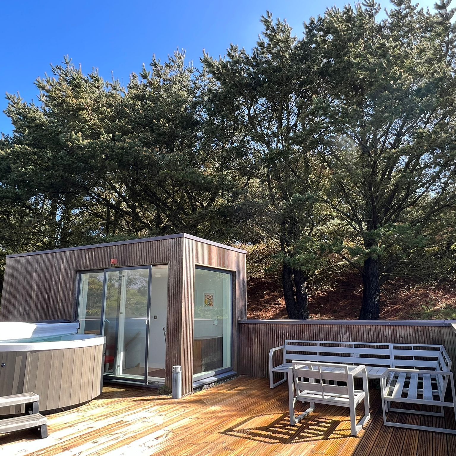 Outdoor wooden deck with a hot tub, lounge chairs, table and glass sliding door structure, bordered by trees under a clear blue sky.