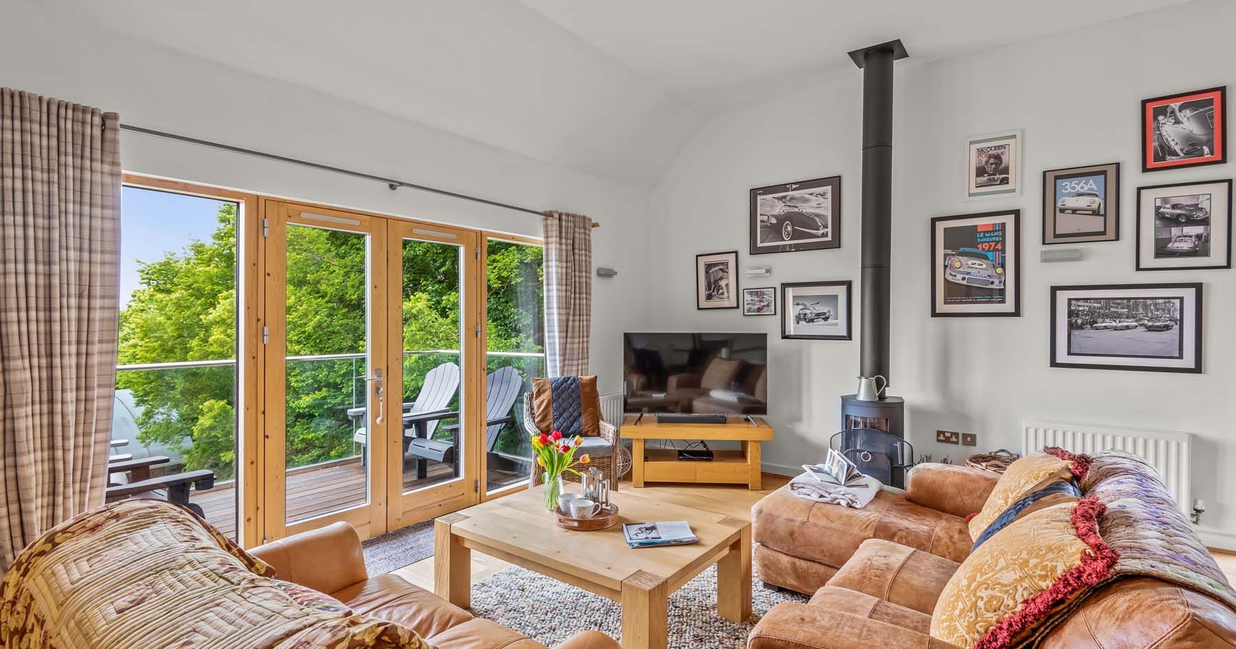 Bright and cozy living room with leather sofas, wood stove, TV, and glass doors opening to a balcony with greenery outside.