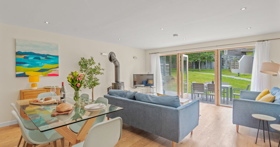 Modern open-plan living and dining room with blue sofas, glass dining table, wood-burning stove, large windows, and doors opening to a patio with outdoor seating and a green lawn.