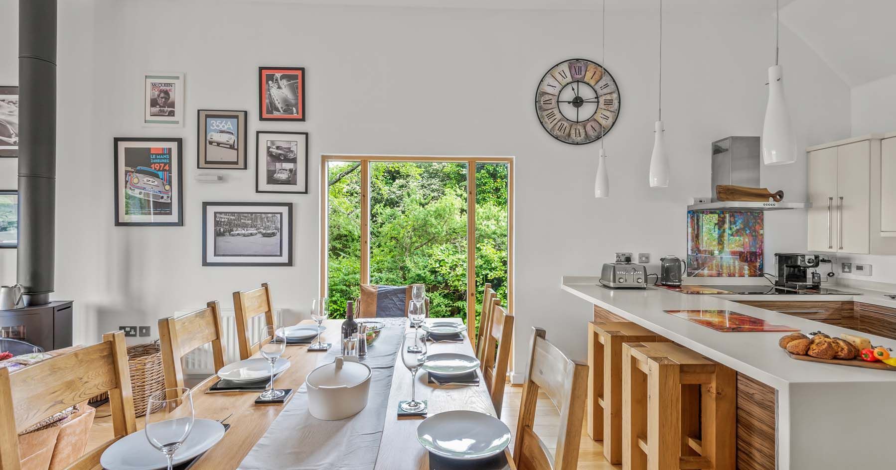 Modern dining room and kitchen with wooden table, white countertops, and large window overlooking greenery.