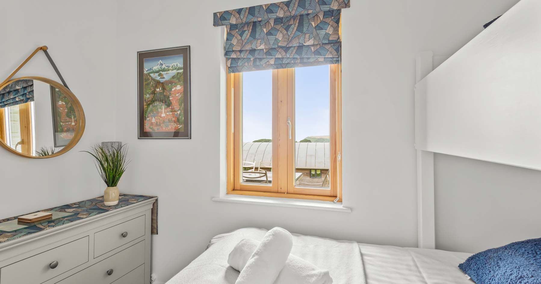 Bright and modern bedroom with window, dresser, mirror, and neatly made bed with towels.