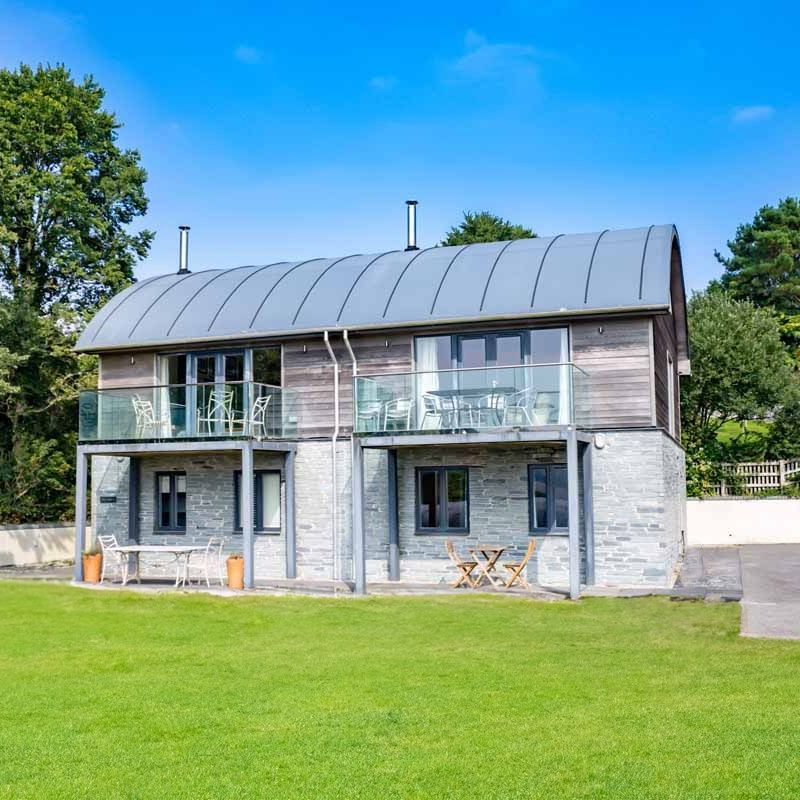Modern two-story house with stone and wood exterior, curved metal roof, glass balconies, and green lawn