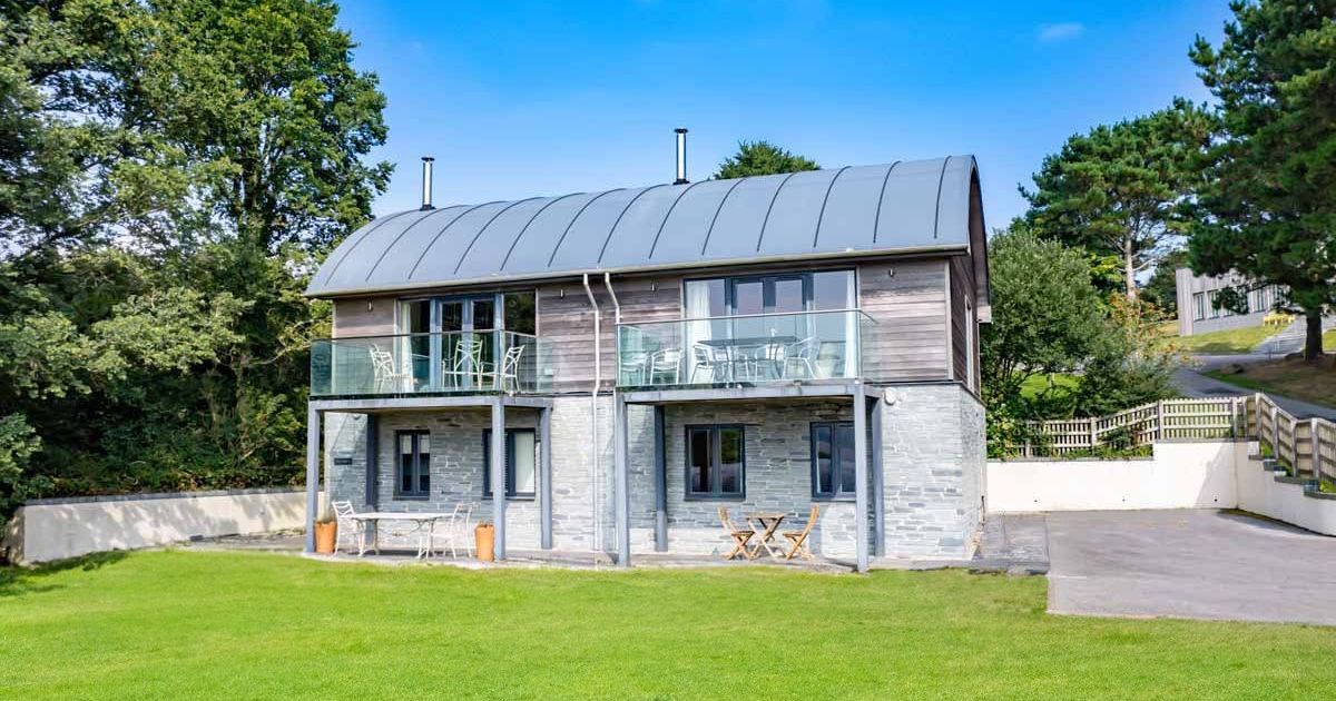 Modern two-story house with stone and wood exterior, curved metal roof, glass balconies, and green lawn