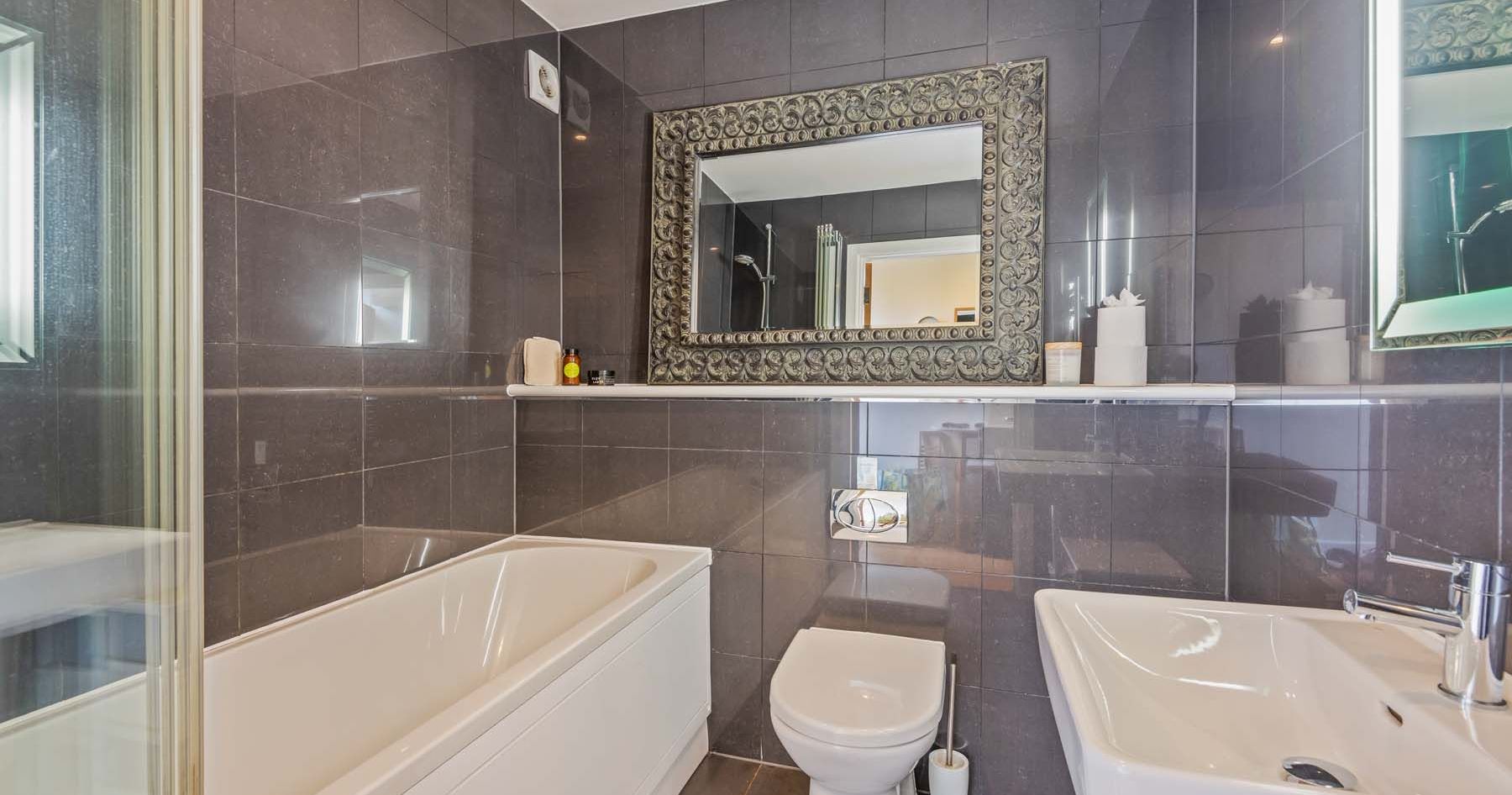Modern bathroom with dark tile walls, bathtub, toilet, sink, and ornate mirror