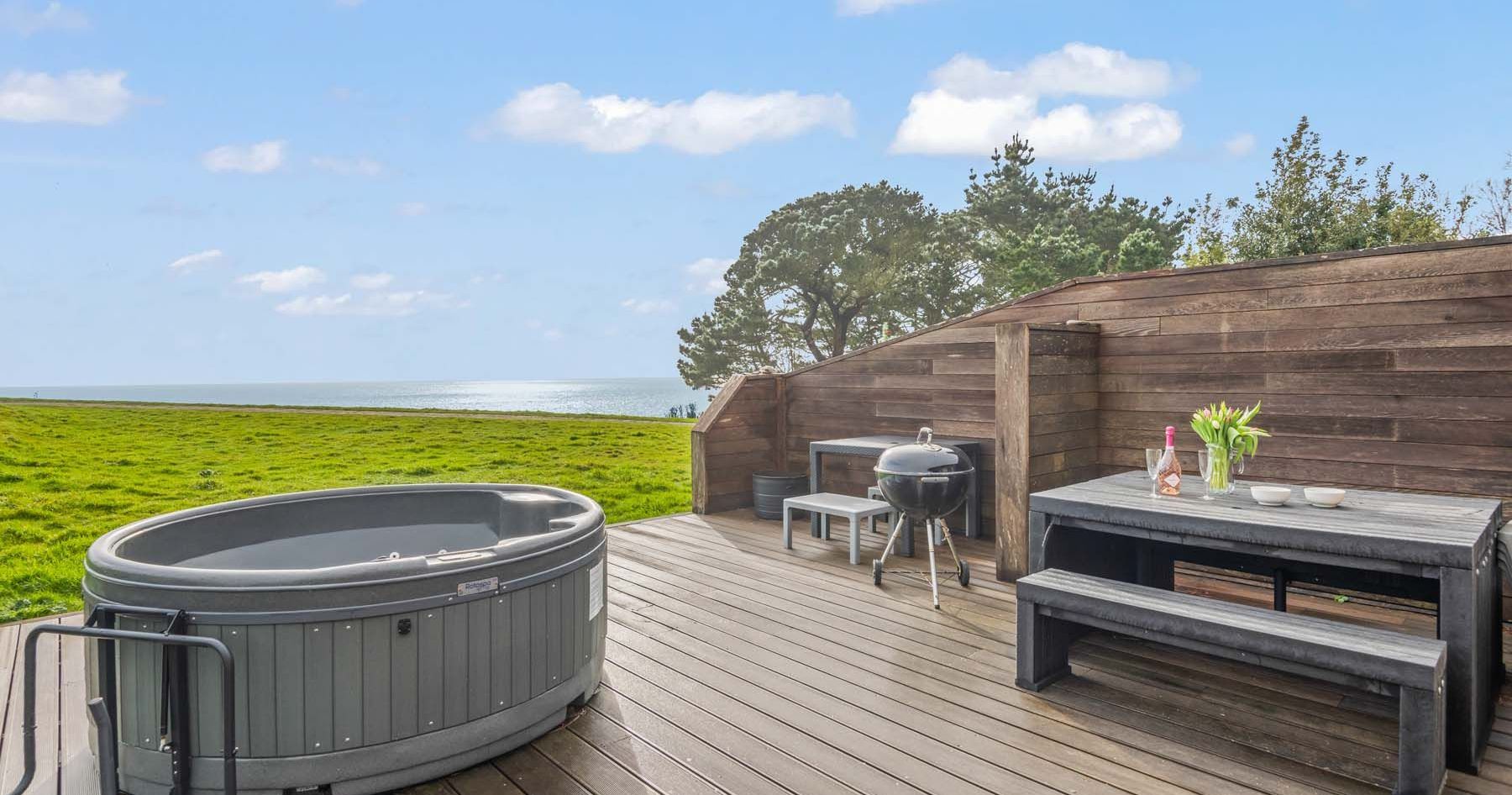 Outdoor deck with hot tub, picnic table, and barbecue overlooking a grassy area and the sea.