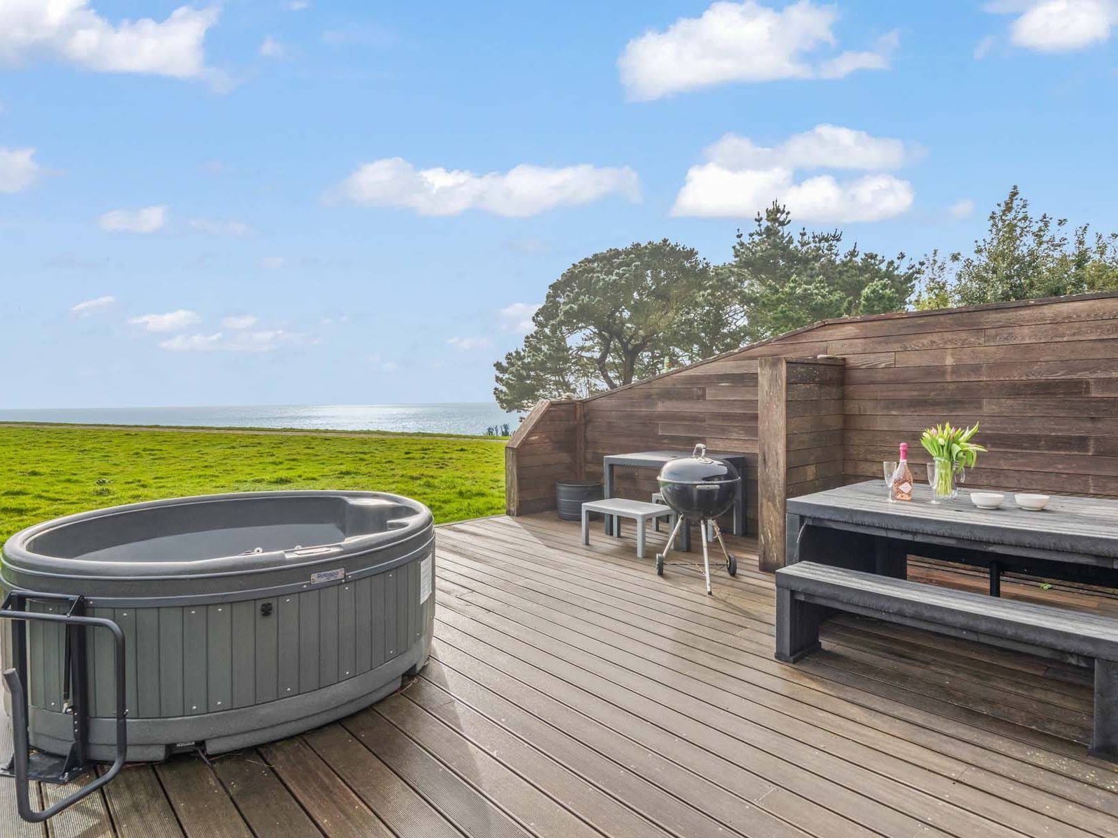 Outdoor deck with hot tub, picnic table, and barbecue overlooking a grassy area and the sea.