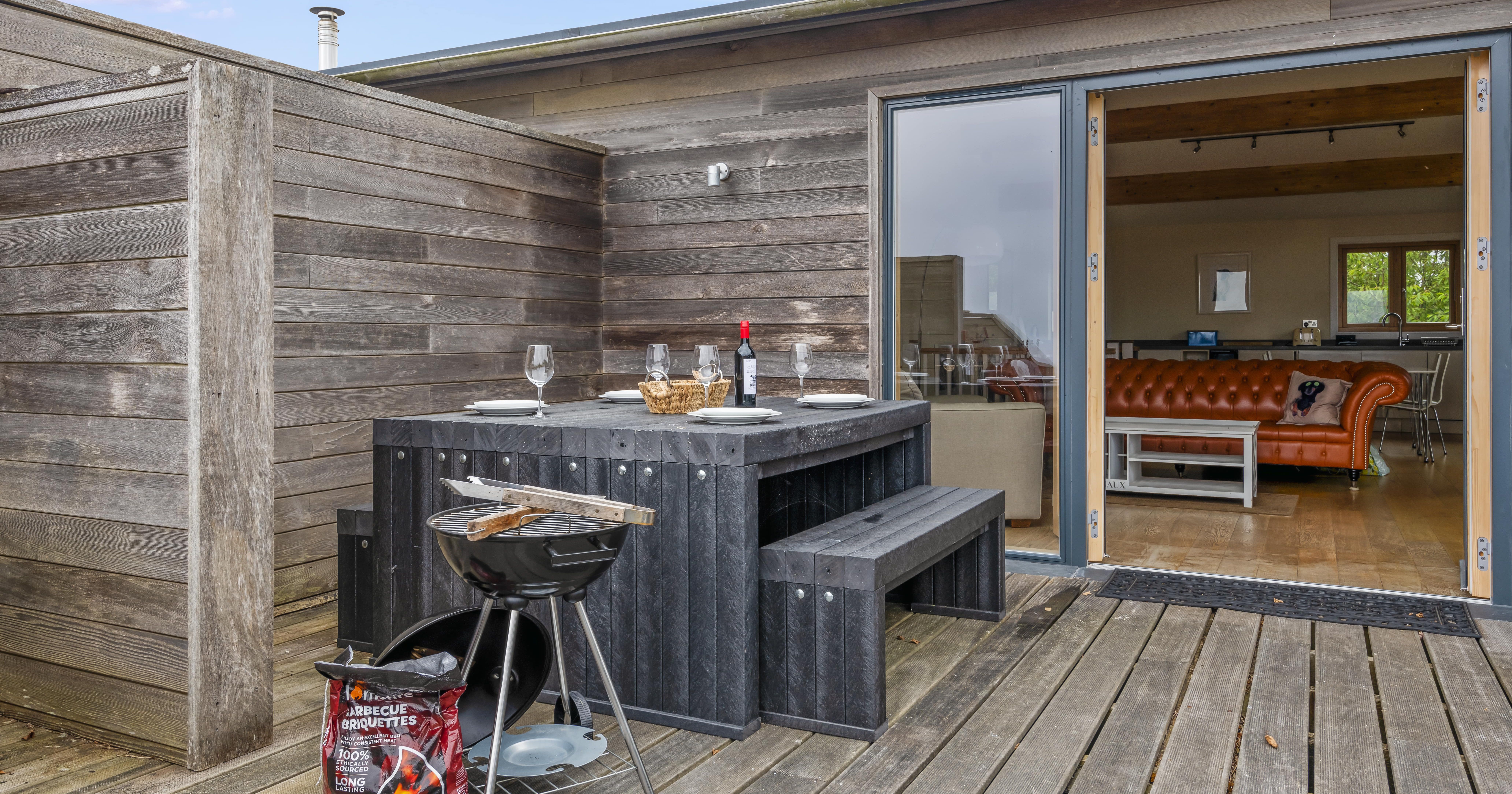 Outdoor patio with wooden table, benches, barbecue grill, and a view into a stylish living room.