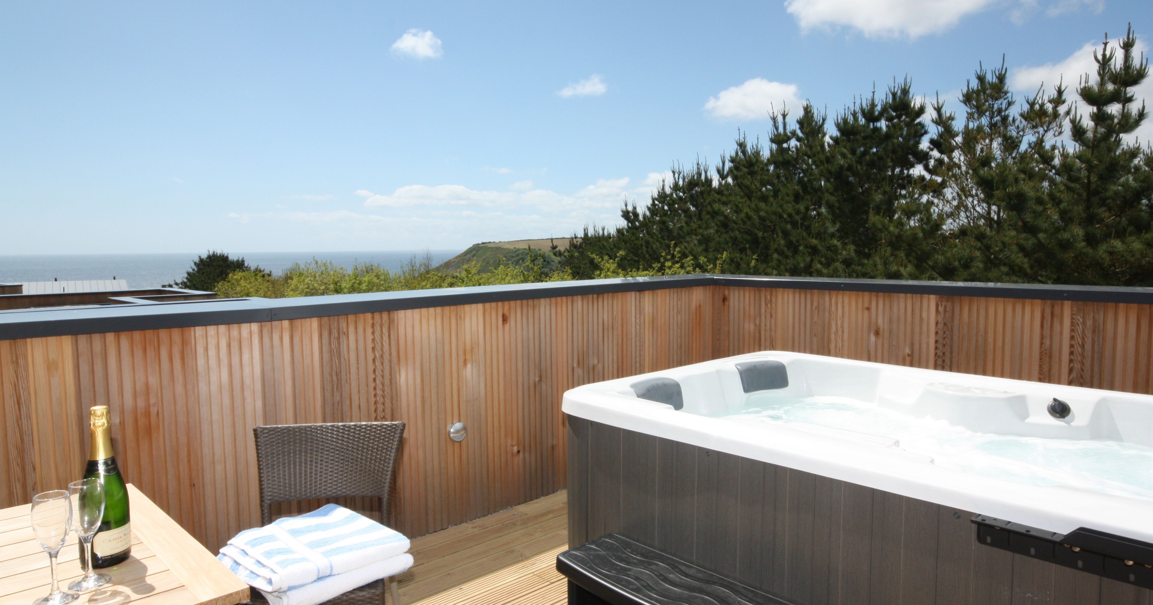 Private outdoor deck with hot tub, wooden fence, table with champagne and glasses, overlooking the sea and trees