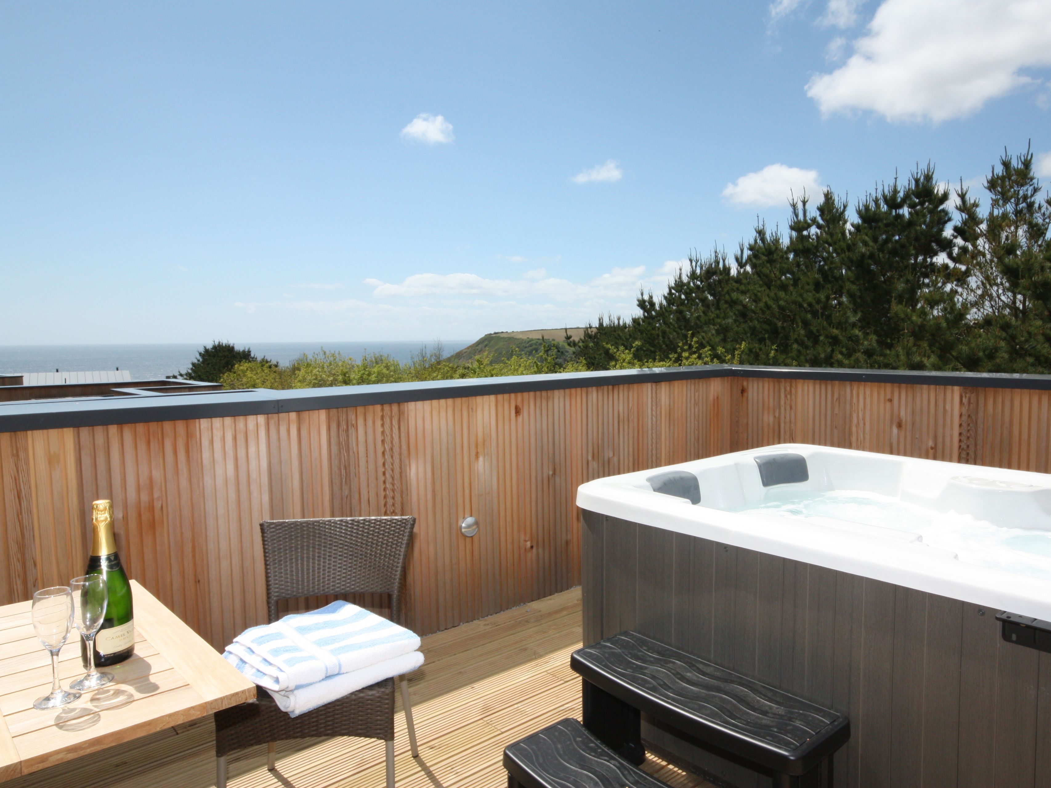 Private outdoor deck with hot tub, wooden fence, table with champagne and glasses, overlooking the sea and trees