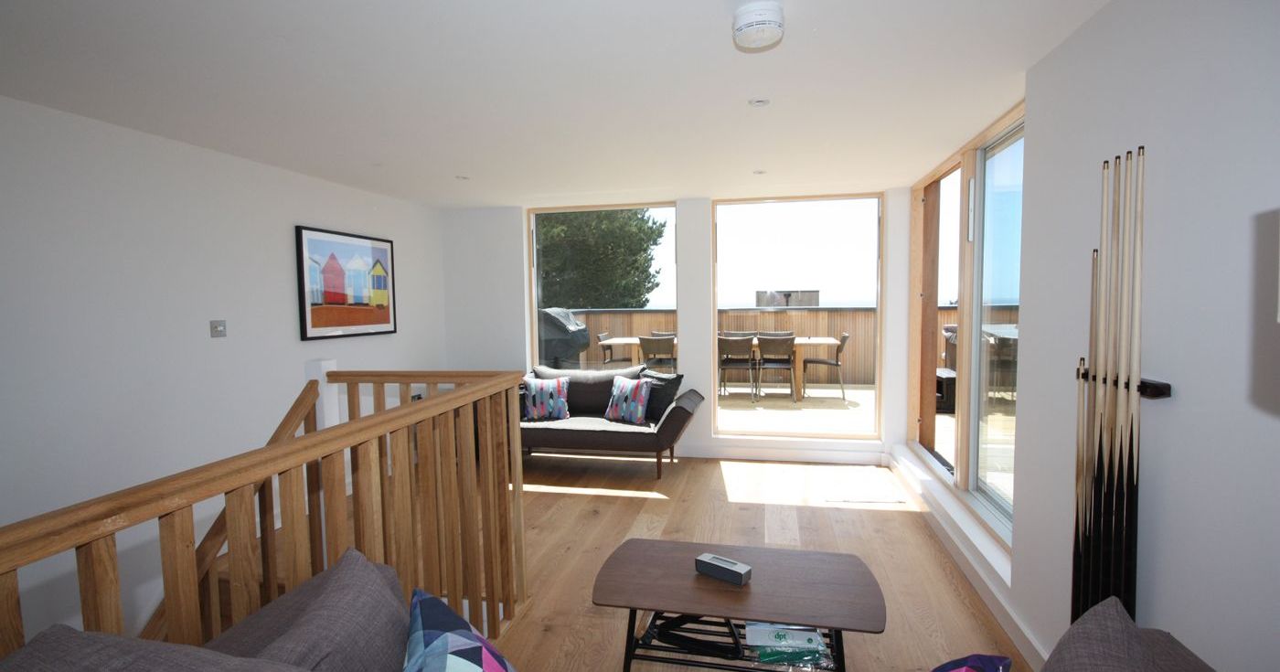 Modern living room with wooden flooring, gray sofas, colorful cushions, large glass doors opening to a patio with outdoor furniture.