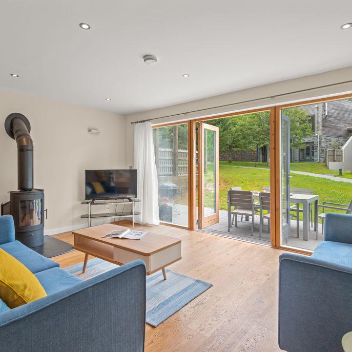 Modern living room with blue sofas and yellow pillows, a wood-burning stove, a flat-screen TV, and sliding glass doors opening to a patio with outdoor dining furniture and a grassy backyard.