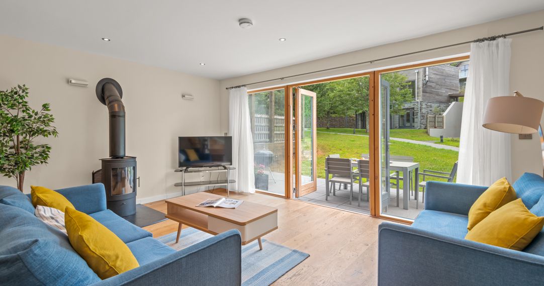 Modern living room with blue sofas and yellow pillows, a wood-burning stove, a flat-screen TV, and sliding glass doors opening to a patio with outdoor dining furniture and a grassy backyard.