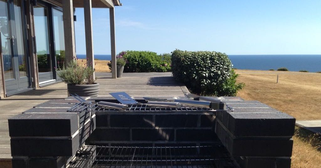 Outdoor brick barbecue grill with utensils, overlooking a grassy area and the sea.