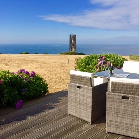 Outdoor patio with wicker furniture overlooking the sea, flowers, and a tall structure in the distance