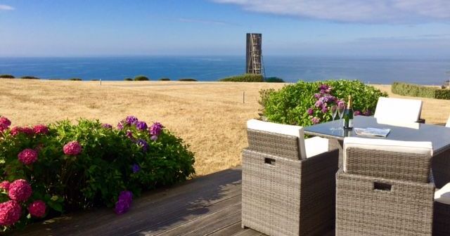 Outdoor patio with wicker furniture overlooking the sea, flowers, and a tall structure in the distance