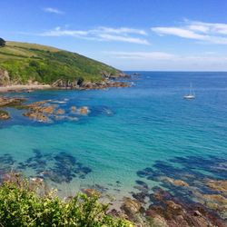 Coastal landscape with turquoise sea, rocky shoreline, green hills, and a sailboat in the distance.