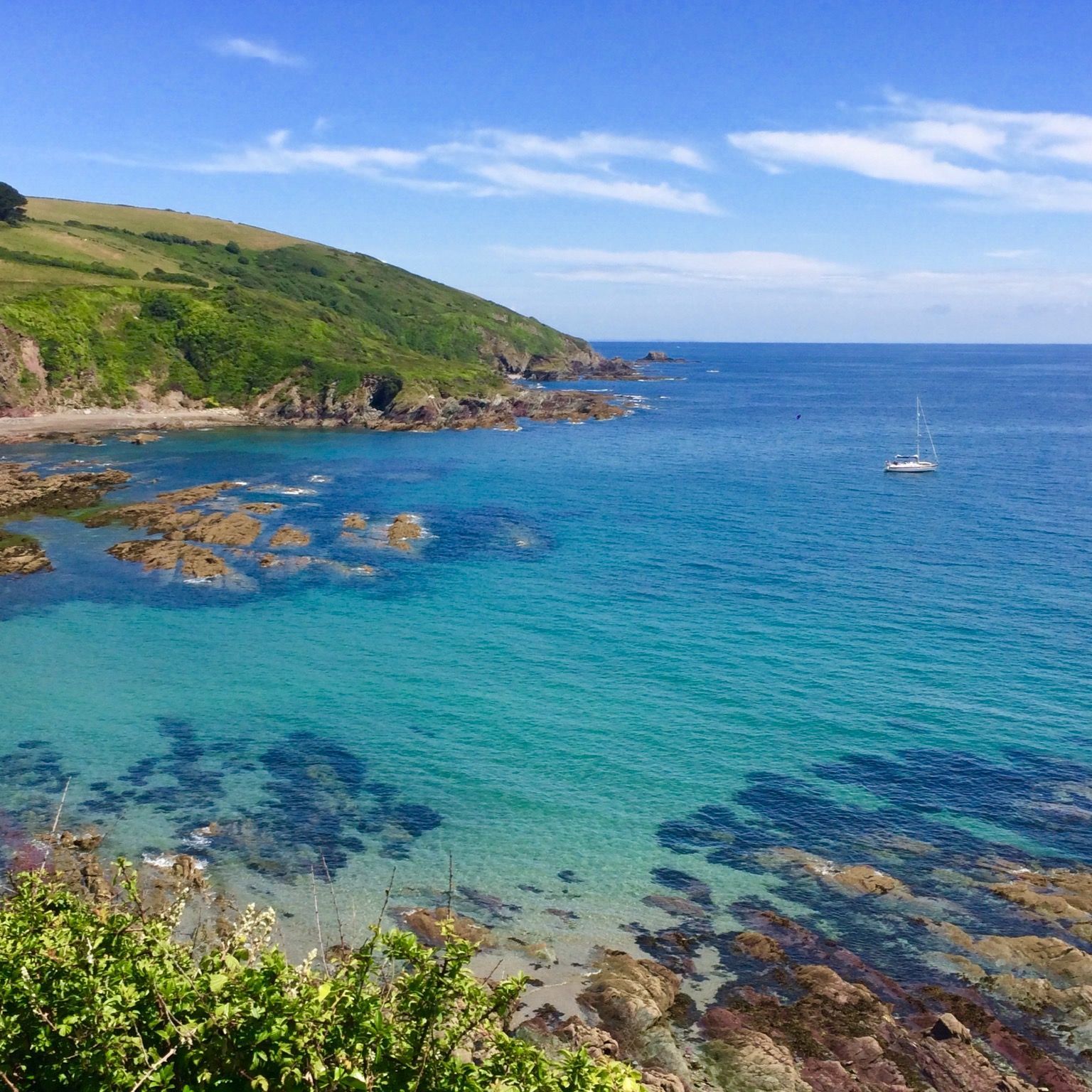 Coastal landscape with turquoise sea, rocky shoreline, green hills, and a sailboat in the distance.