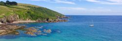 Coastal landscape with turquoise sea, rocky shoreline, green hills, and a sailboat in the distance.