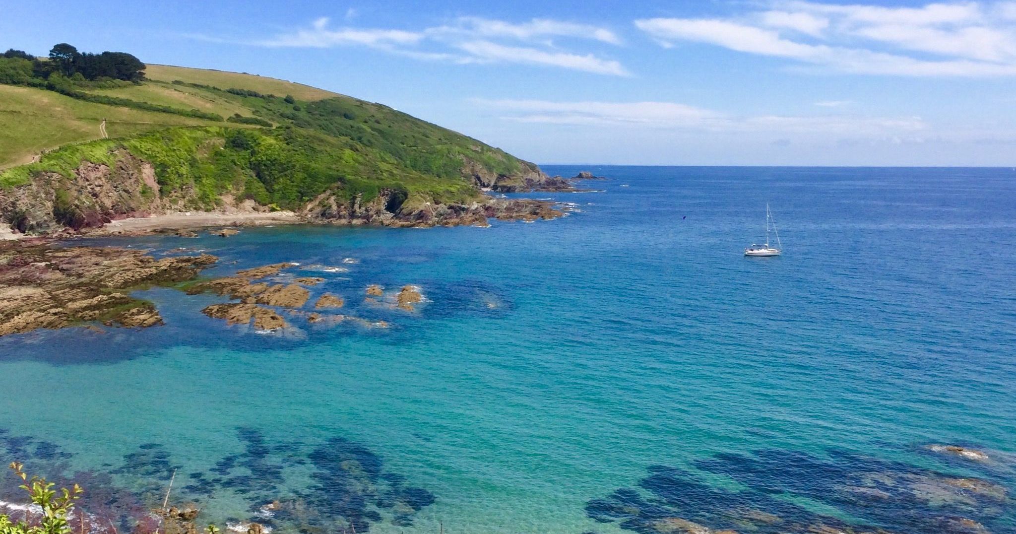 Coastal landscape with turquoise sea, rocky shoreline, green hills, and a sailboat in the distance.