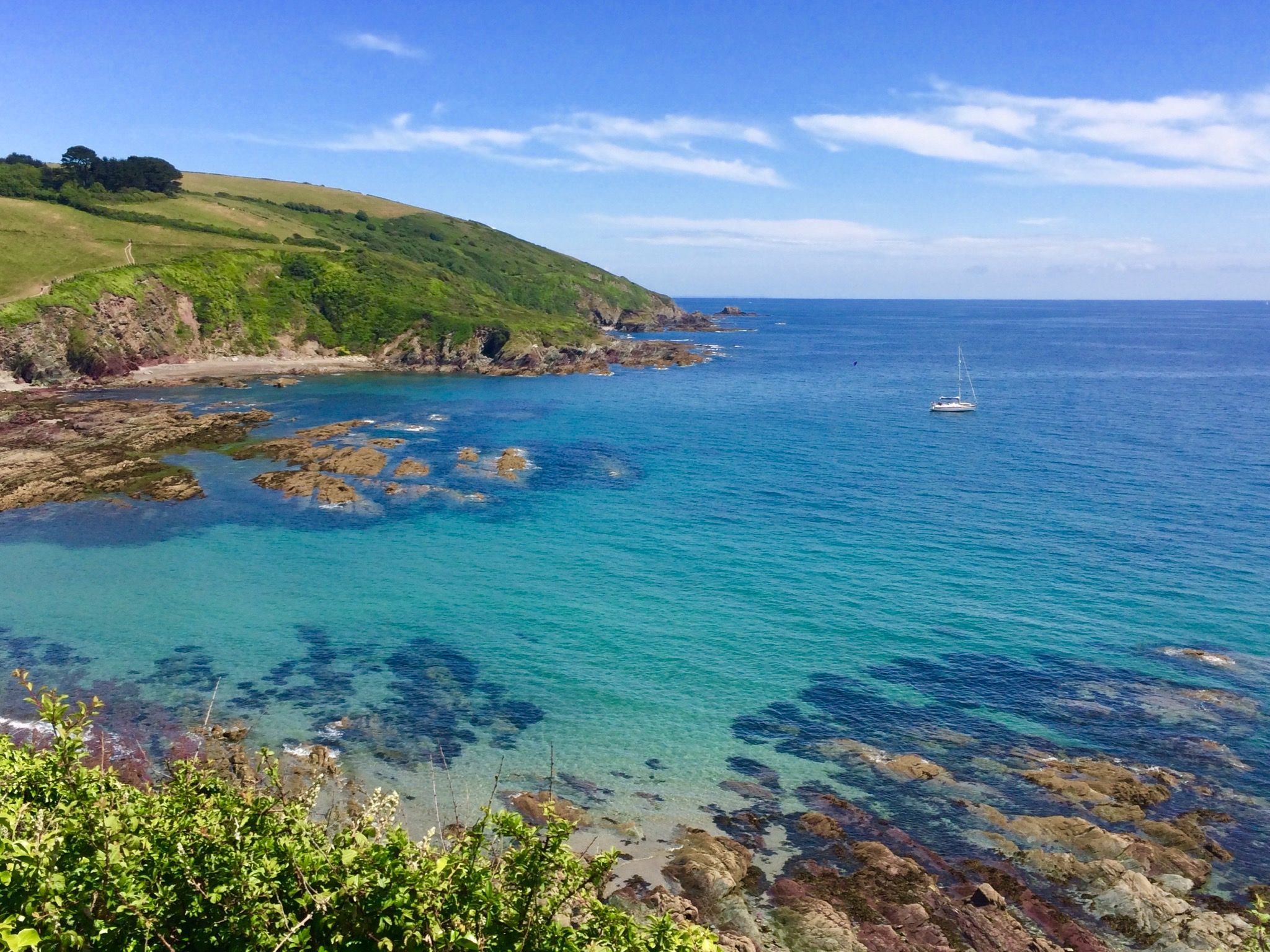 Coastal landscape with turquoise sea, rocky shoreline, green hills, and a sailboat in the distance.