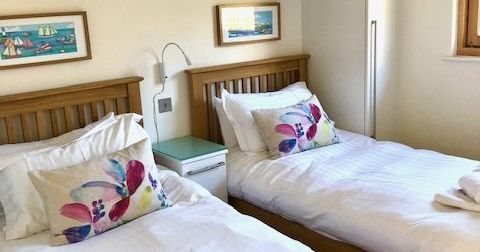 A bright bedroom with two single beds, each with floral pillows, white linens, and neatly folded towels.