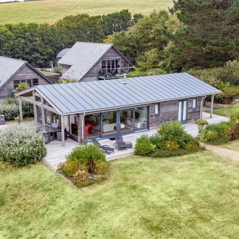Aerial view of a modern wooden house with large windows and a spacious deck, surrounded by greenery and other buildings.