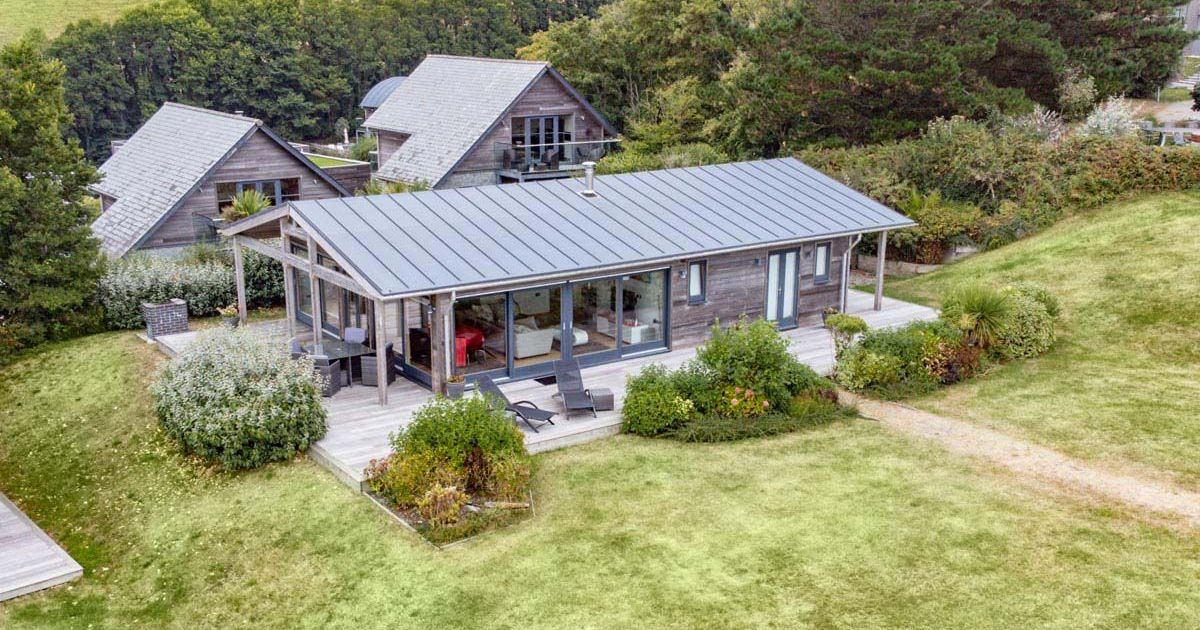 Aerial view of a modern wooden house with large windows and a spacious deck, surrounded by greenery and other buildings.