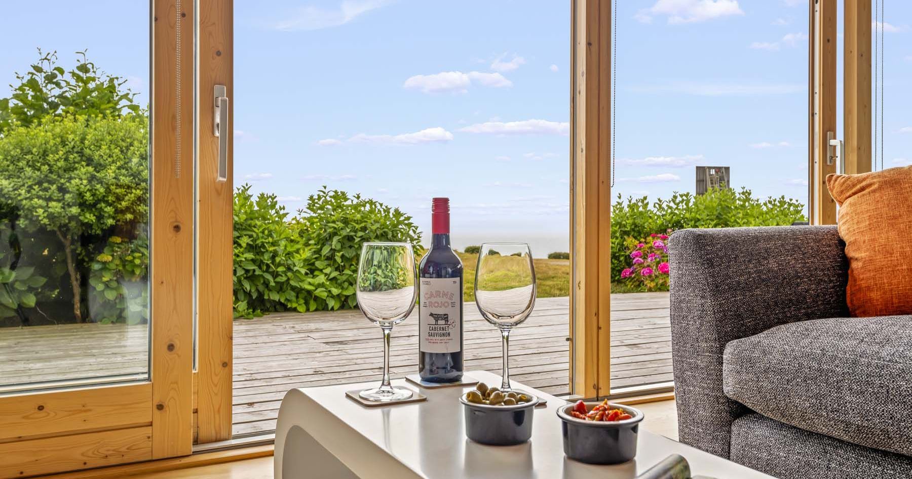Modern living room with large glass doors overlooking a wooden deck, wine, glasses, and snacks on a table.