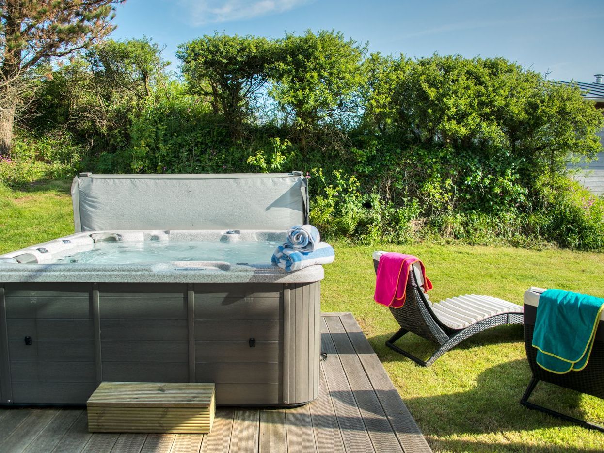 Outdoor hot tub with rolled towels next to two lounge chairs with colorful towels in a grassy garden.