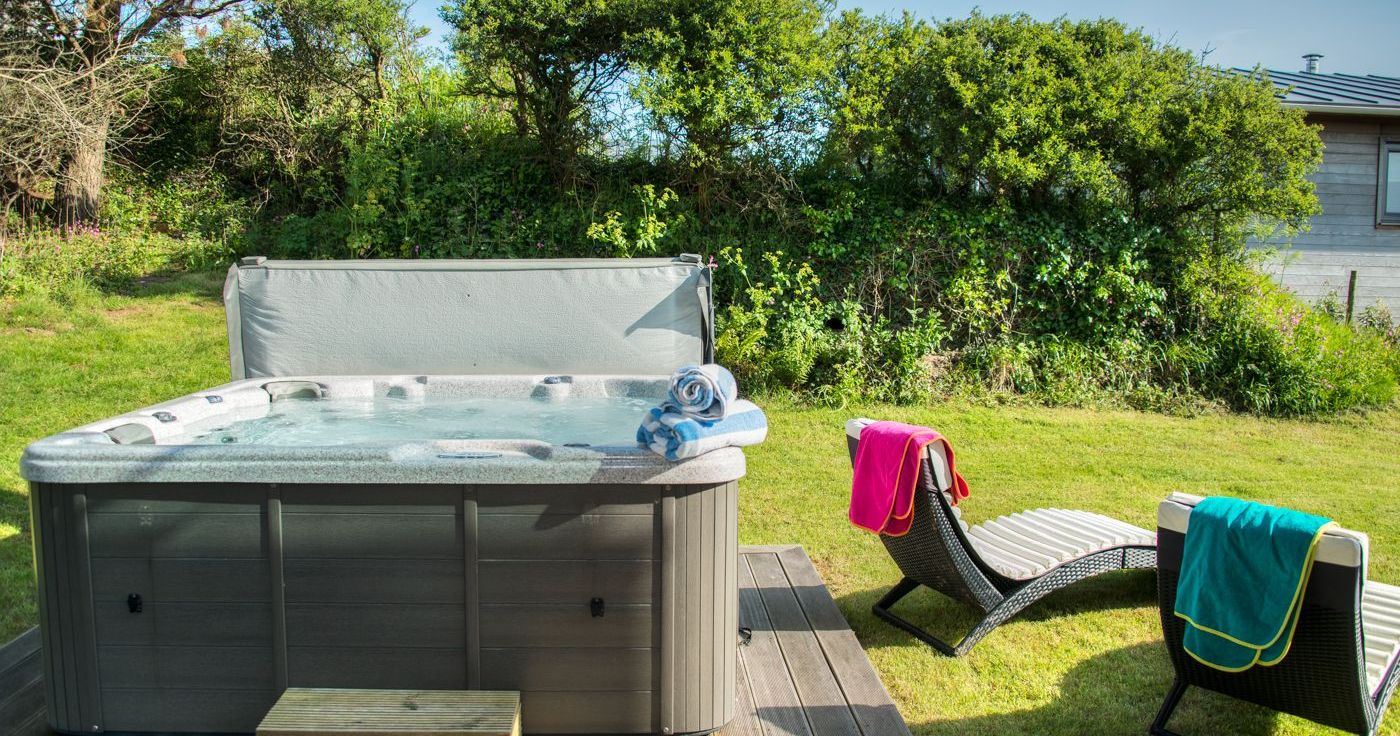 Outdoor hot tub with rolled towels next to two lounge chairs with colorful towels in a grassy garden.