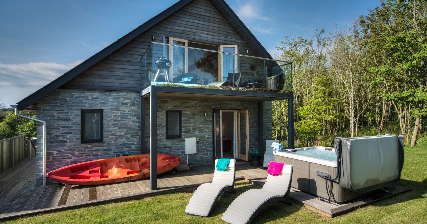 Modern house with stone and wood exterior, upper balcony, kayak, hot tub, and lounge chairs on a grassy lawn.