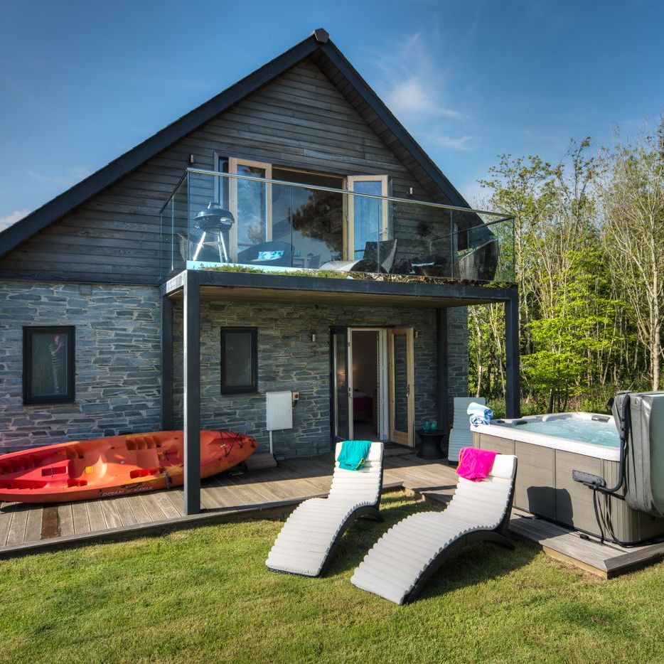 Modern house with stone and wood exterior, upper balcony, kayak, hot tub, and lounge chairs on a grassy lawn.