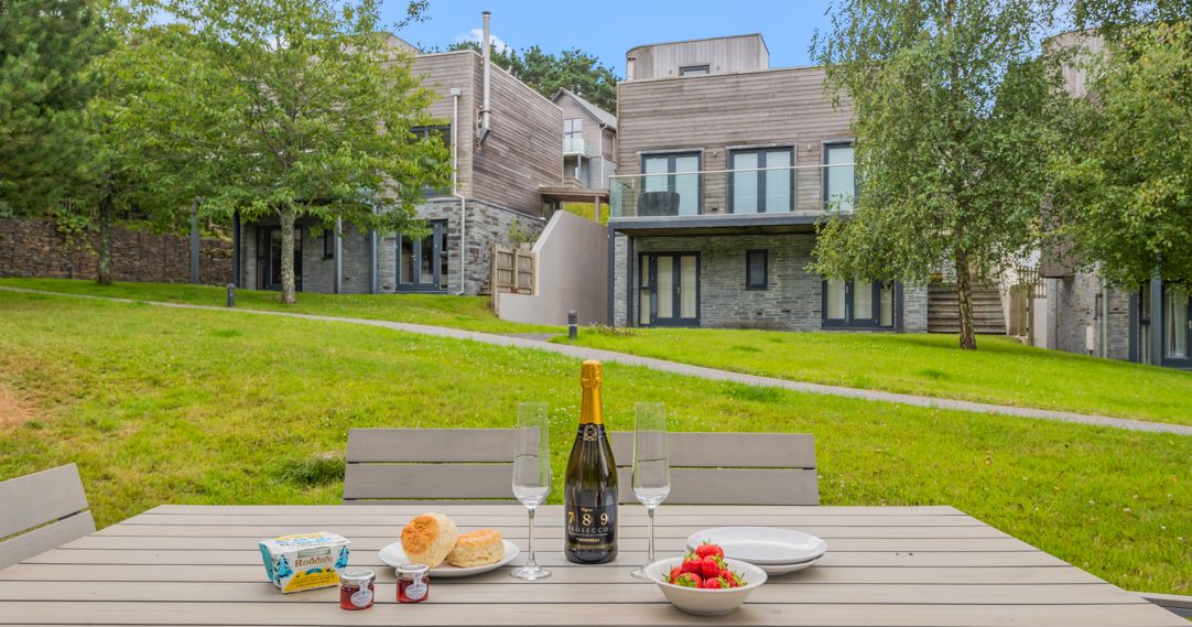 Outdoor table set with champagne, scones, jam, butter, and strawberries, with modern wooden cottages and green grassy area in the background.