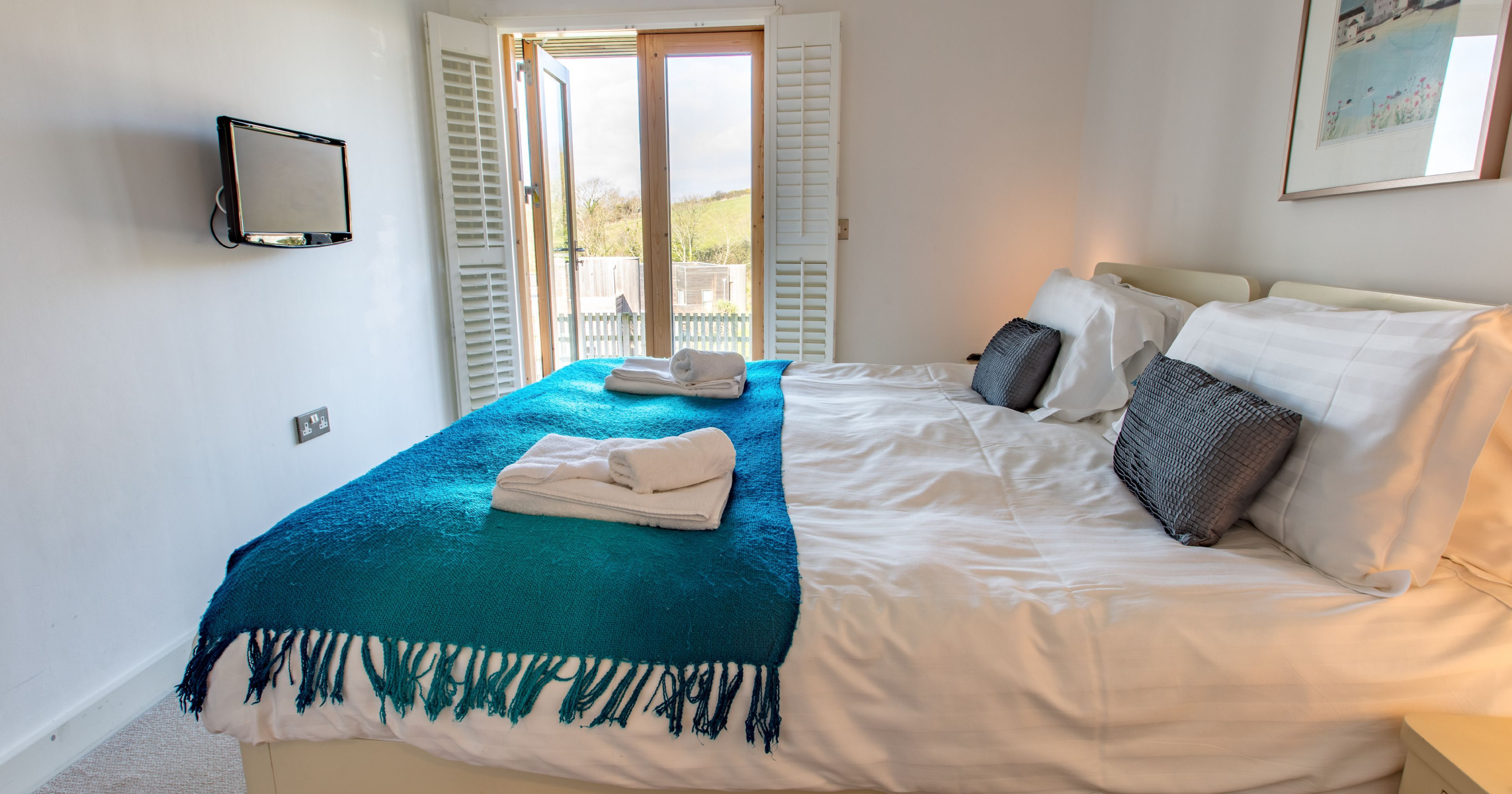 Bright bedroom with large bed, teal throw blanket, pillows, and double doors opening to a view outside.