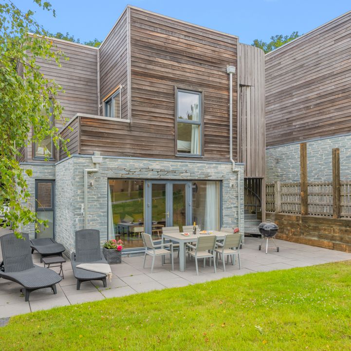 Modern townhouse with wooden and stone exterior, outdoor dining area, sun loungers, patio, and grassy garden.