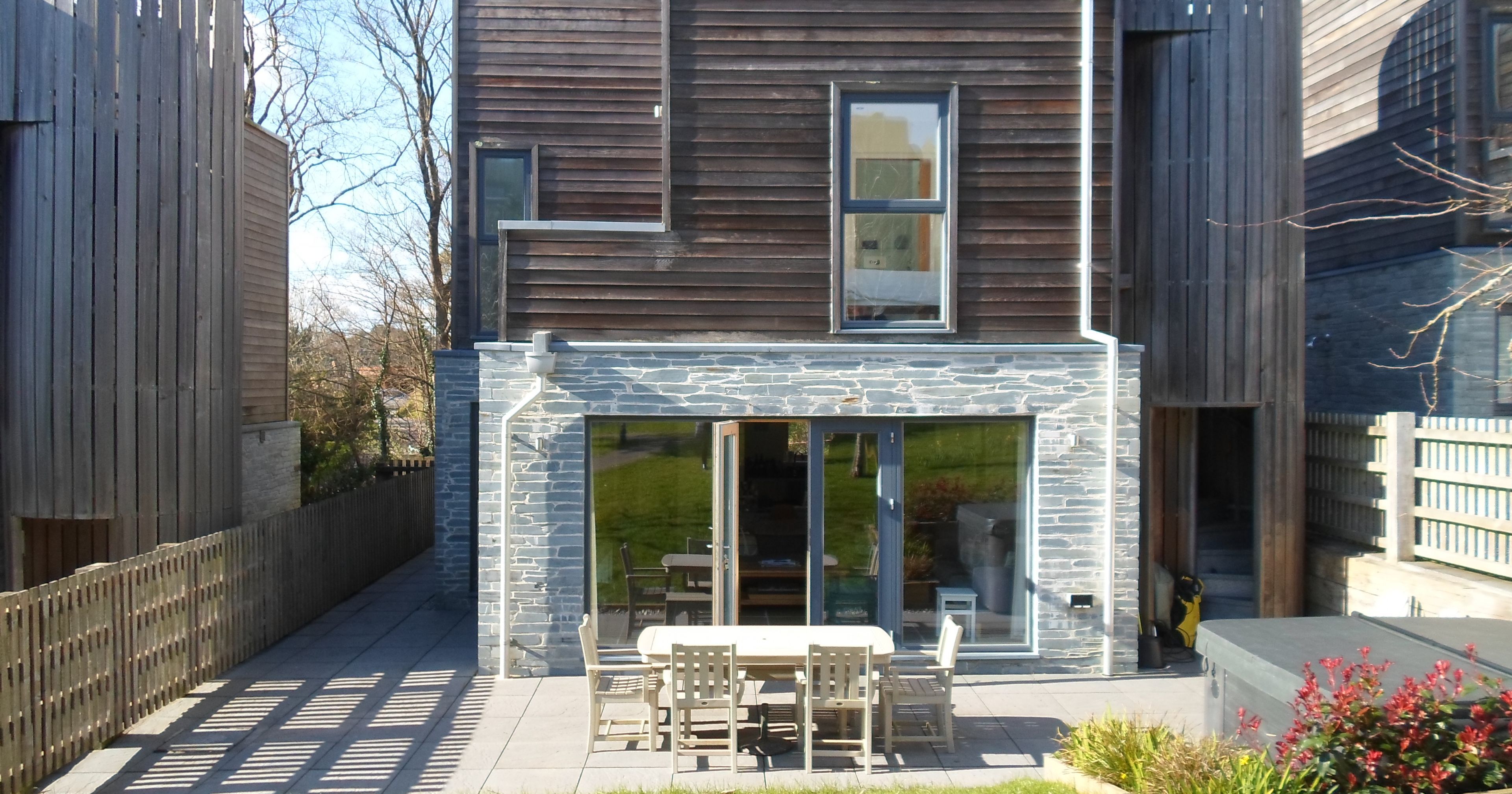 Modern two-story house with wooden and stone exterior, patio area with outdoor dining set, and a grassy backyard.