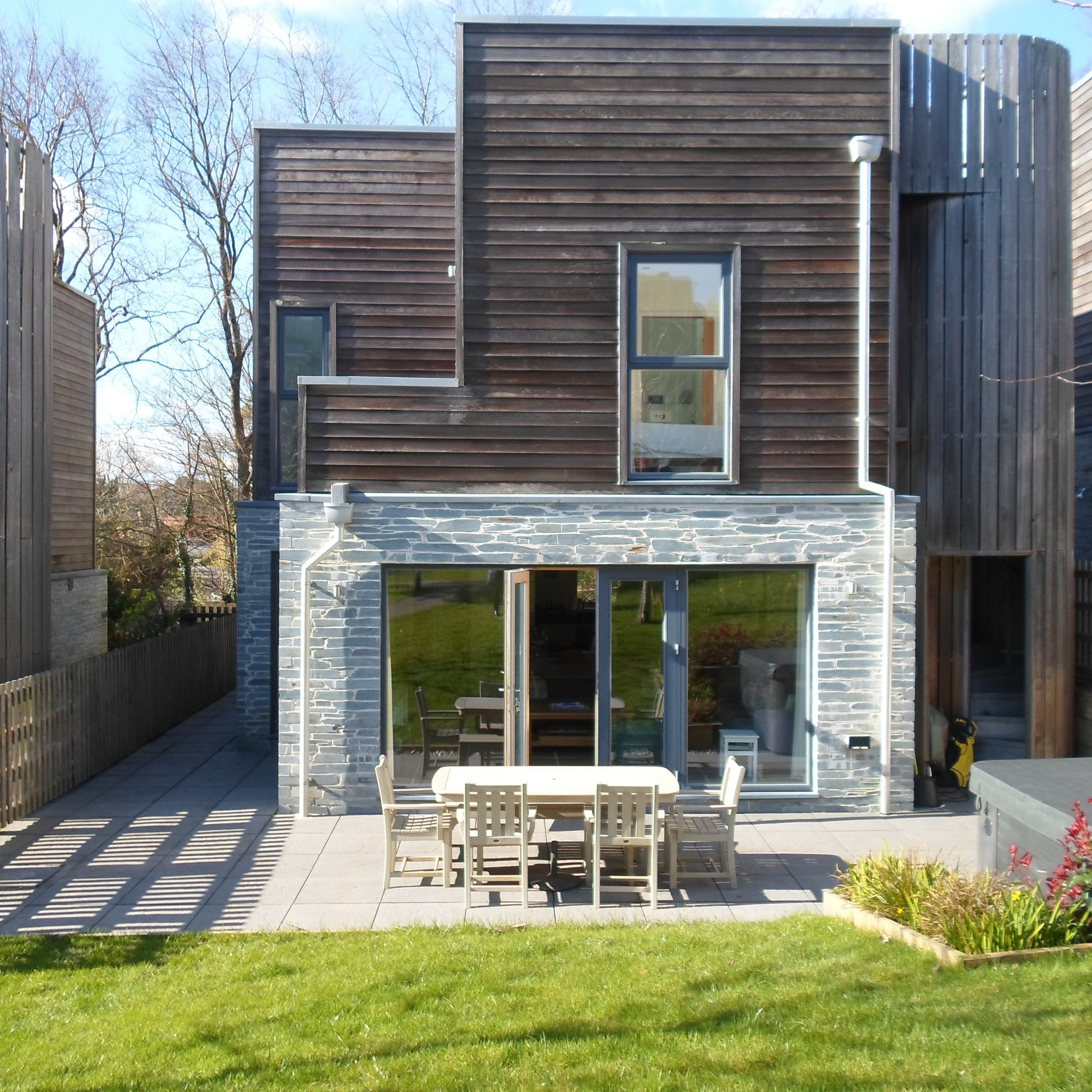 Modern two-story house with wooden and stone exterior, patio area with outdoor dining set, and a grassy backyard.